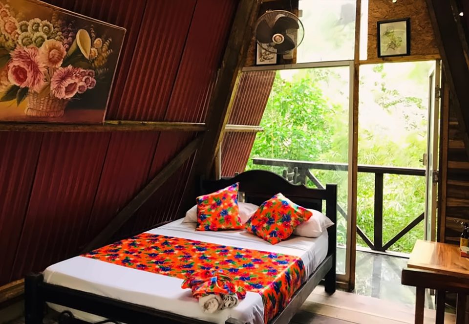Cama doble con colcha y cojines de flores naranjas en una cabaña A-frame, toallas dobladas, ventilador de pared, un cuadro de flores y balcón de madera hacia la selva.