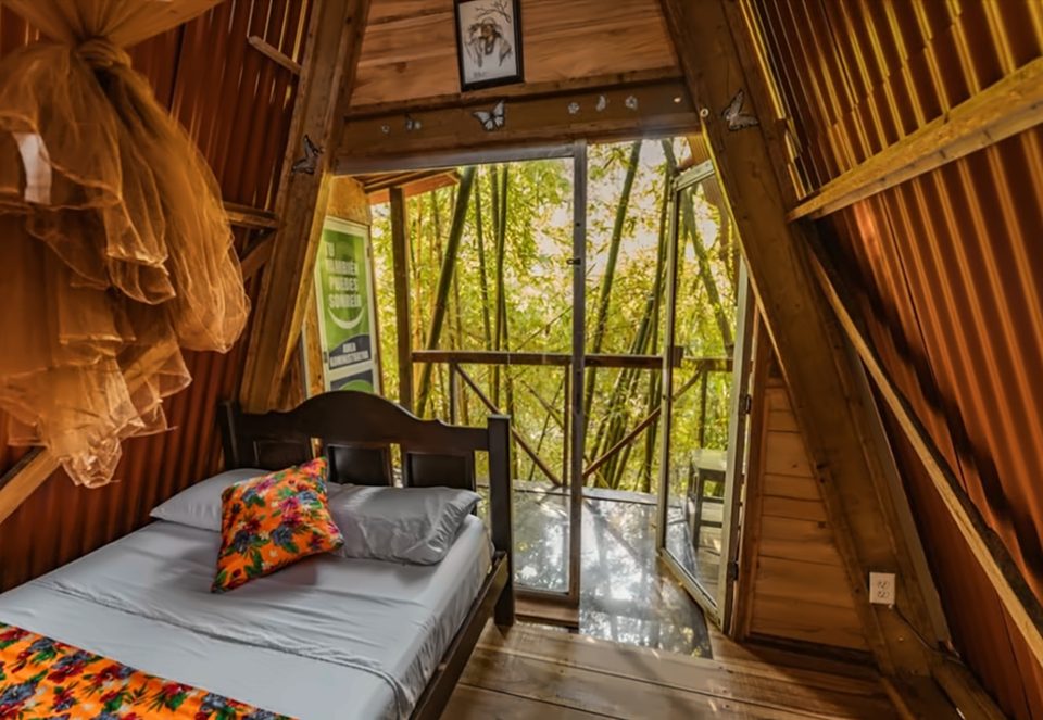 Interior de una cabaña A-frame: cama doble con colcha de flores, toldillo recogido, techo triangular de madera y lámina y puerta de vidrio abierta hacia un bosque de bambú.