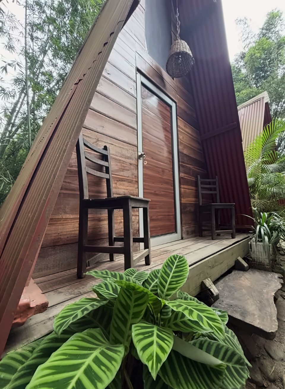 Porche de una cabaña A-frame con dos sillas de madera, puerta de madera, una lámpara de mimbre colgante y una calatea rayada en primer plano.
