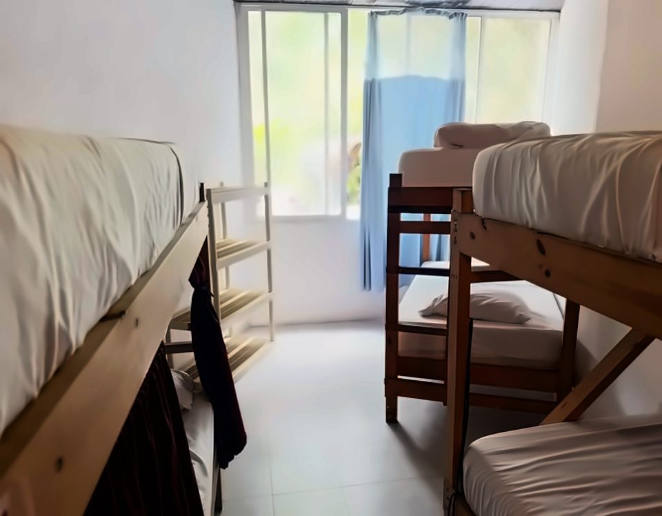Dormitorio compartido con camarotes de madera y sábanas blancas, una repisa abierta y una ventana grande con cortina azul.