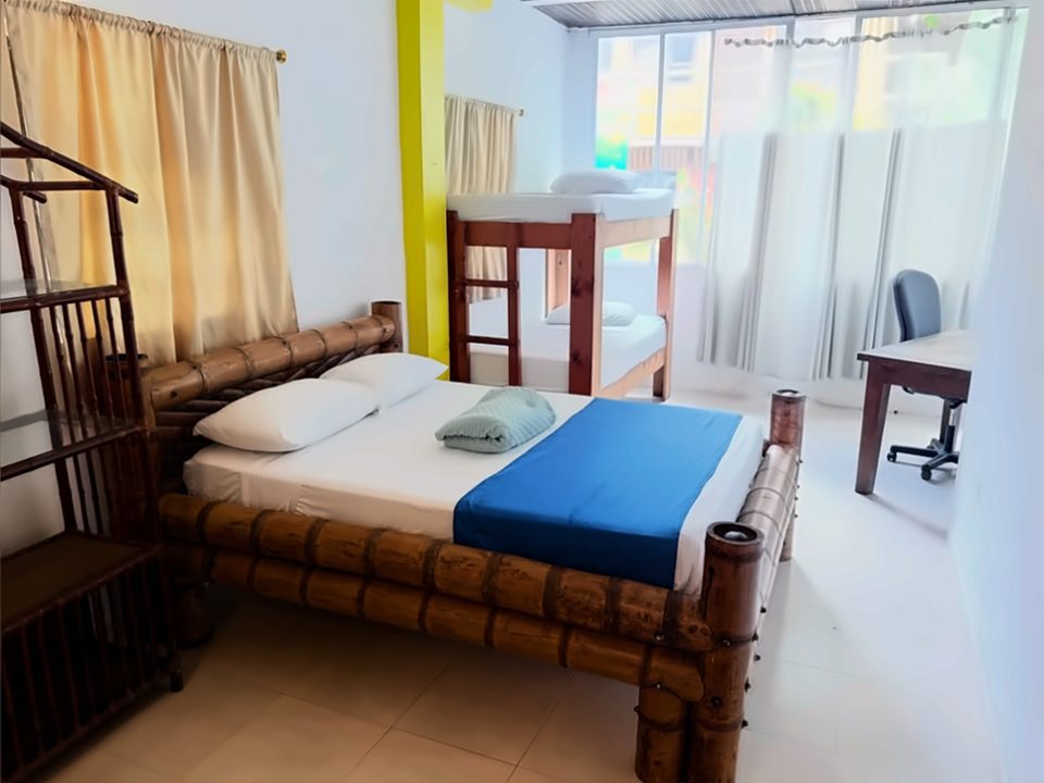 Habitación familiar con cama doble de bambú y cobija azul, un camarote de madera al fondo, escritorio con silla y ventanal con cortinas.