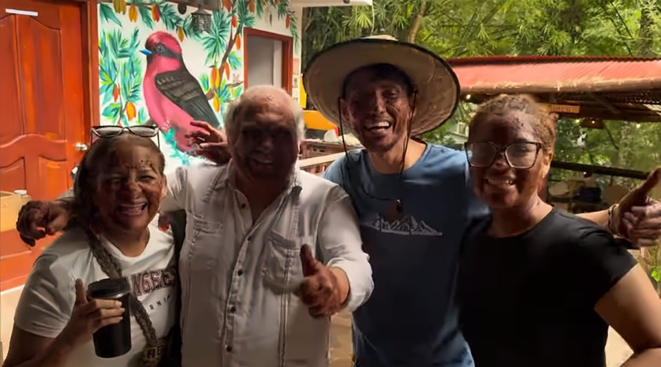 Cuatro personas sonrientes posan abrazadas, dos con el pulgar arriba, frente a un mural de un pájaro rojo entre hojas y frutos.